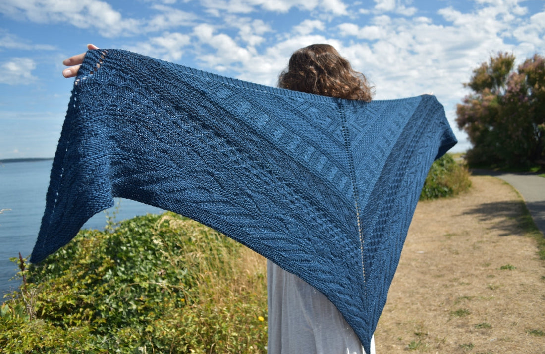 Person outdoors holding a large, textured blue knitted shawl with scenic water and sky background